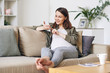 © pressmaster - Smiling beautiful young pregnant woman in casual shirt sitting on sofa and using smartphone while reading internet articles about baby-raising