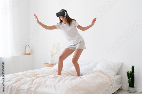 Young female standing on bed, wearing virtual reality headset, spreading arms as if flying in game, smiling and enjoying her VR activity