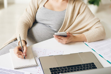 Canvas Print - Close-up of unrecognizable pregnant businesswoman with smartphone in hand sitting at table and making notes about tasks while viewing data on laptop