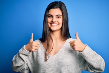 Young beautiful brunette woman wearing casual sweater standing over blue background success sign doing positive gesture with hand, thumbs up smiling and happy. Cheerful expression and winner gesture.