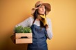 © Krakenimages.com - Young beautiful brunette gardener woman wearing apron and hat holding box with plants serious face thinking about question, very confused idea