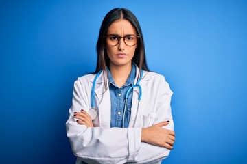 Wall Mural - Young beautiful doctor woman wearing stethoscope and glasses over blue background skeptic and nervous, disapproving expression on face with crossed arms. Negative person.