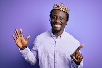 Young african american man wearing golden crown of king over isolated purple background showing and pointing up with fingers number six while smiling confident and happy.