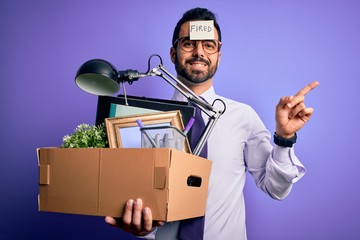 Poster - Young handsome businessman with beard holding cardboard box with fired reminder on head very happy pointing with hand and finger to the side