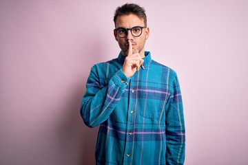 Young handsome man wearing casual shirt and glasses standing over isolated pink background asking to be quiet with finger on lips. Silence and secret concept.