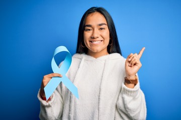 Canvas Print - Young asian woman holding blue cancer ribbon symbol standing over isolated background very happy pointing with hand and finger to the side