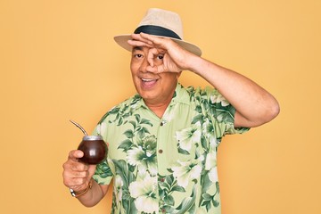 Canvas Print - Middle age senior grey-haired man wearing summer hat drinking traditional mate drink with happy face smiling doing ok sign with hand on eye looking through fingers