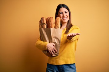 Sticker - Young beautiful woman holding a bag of fresh healthy bread over yellow background smiling cheerful offering palm hand giving assistance and acceptance.