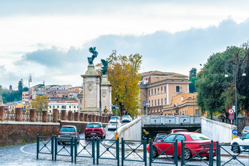 Rome, Italy. Vehicular traffic on cobblestone streets of the city. Statues of angels with wreaths and sword, underground road/ roadway, Roman architecture buildings in background.