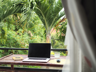 laptop of a remote digital nomad on a wooden bamboo table with notebook, mobile phone and glass in nature on a balcony with a green tropical background with palm trees