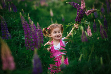 A small cheerful girl with two light tails on her head in a green field with purple flowers. A girl in a pink dress in a lupine field and a bouquet of lupines in her hands. Child's emotions-joy, prank