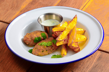 Two meat cutlets with baked potato and sauce in a saucepan. Served in a white plate over dark rustic wooden background.