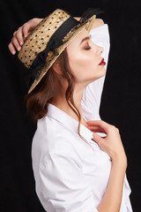 A brown haired lady in a white shirt and a straw hat with a black ribbon, a bow-knot and covered with mesh cloth is posing on the black backdrop. The lady is holding the fashion hat on her head. 