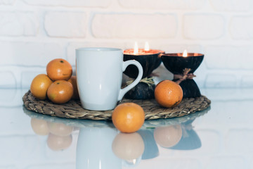 Wall Mural - Burning spa aroma candles in coconut shell, cup of tea, tangerines on wicker stand on a glass white table, cozy home interior background.