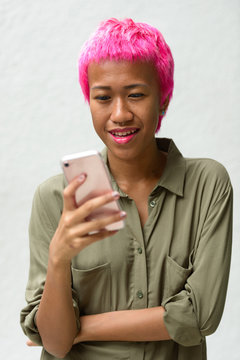 Happy young rebellious Asian woman with pink hair using phone