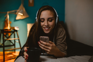 woman laying bed at her room using mobile phone wearing headphones