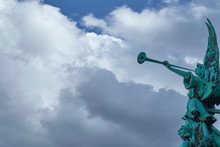 Trumpeting Old Bronze Angel Statue Covered With Green Verdigris Playing A Celestial Fanfare Against A Sunny Sky With Dramatic Clouds.