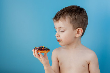 Wall Mural - Little boy eating donut chocolate on blue background. Cute happy boy smeared with chocolate around his mouth. Child concept, tasty food for kids