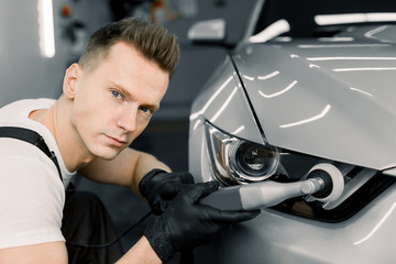 Car detailing series, polishing concept. Professional male auto service worker, wearing white t-shirt and overalls, waxing and polishing headlight of white car with polish machine, looking at camera