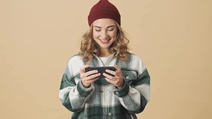 Canvas Print - Cheerful pretty woman in knit hat and shirt playing on smartphone over beige background