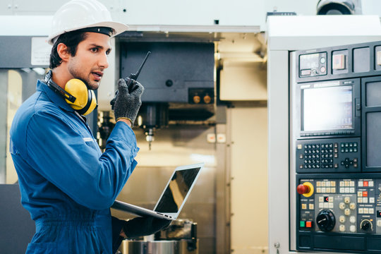 Male industrial engineer or technician in safety hard helmet and uniform using laptop checking on machine and radio phone calling team worker, heavy technology invention industry manufacturing factory