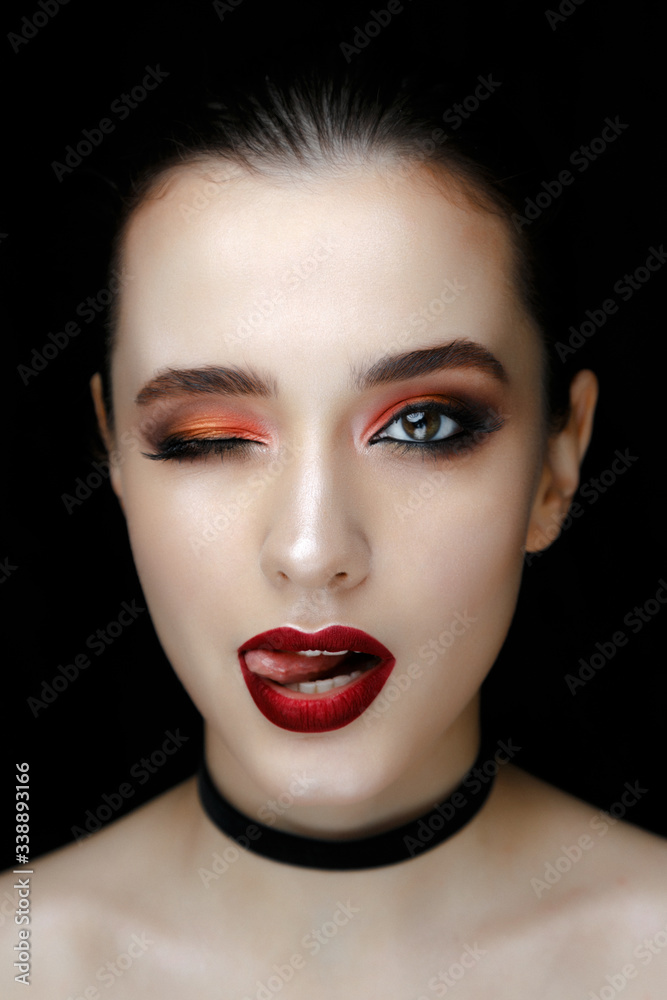 Close-up portrait of gorgeous young woman with red lips and bright makeup of eyes looks to the camera.. Beautiful girl with a healthy clean skin against black background. - obrazy, fototapety, plakaty 