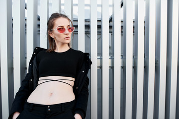 Young woman with long hair in pink sunglasses in a black fashionable top in a light summer jacket standing near a gray metal wall. Urban girl relaxed.Street style