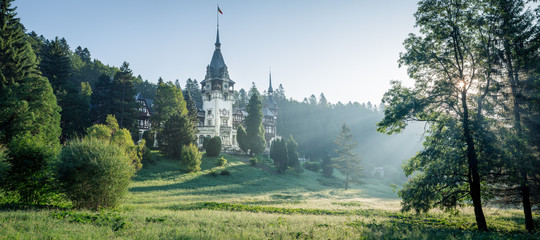 Peles Castle, famous residence of King Charles I in Sinaia, Romania. Peaceful summer landscape at sunrise of royal palace and park with mist and sun rays shining through the branches.