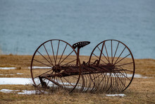 A Vintage Rusty Hay Rake Sitting In A Meadow With Yellow Grass And Patches Of Snow. The Farm Rake Has A Round Rusty Seat And Is A Horse-drawn Farm Tool. There's An Ocean In The Background.