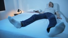 A Man Is Lying In A Bed Watching Tv, Operating A Remote Control And Yawning Big, Room Illuminated By Tv Flicker