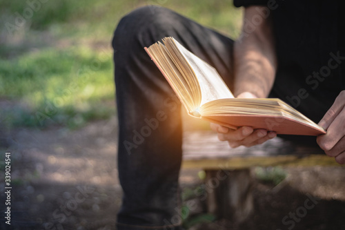 Man sitting on a bench and reading a book outside in the park. Male hands holding a book outdoor. Student studying in nature, focus on the book, copy space for text.