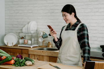 Young asian japanese woman with fresh vegetables smiling while using mobile phone in modern kitchen. beautiful lady in apron text message online on cellphone during cooking time with healthy meal.