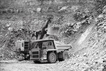 Excavator and dump truck while loading stone ore in a quarry, black and white. Mining industry.