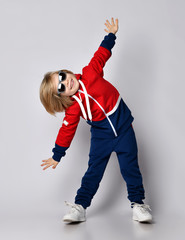 Wall Mural - Happy smiling active blond kid boy in blue and red hoodie, pants and sunglasses does gym exercises