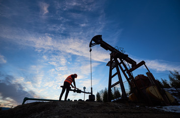 Silhouette of oil man in work vest and helmet. Petroleum operator working in oil field with long stroke balanced beam petroleum pump jack under beautiful sky. Concept of petroleum industry.