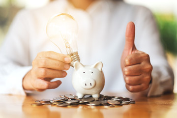 A woman making and showing thumbs up hand sign while putting light bulb over a piggy bank on the table for saving money concept