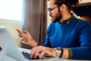 Wall Mural - Attractive Caucasian bearded entrepreneur sitting in his home office, using smart phone and taking a break from work. Work from home concept.