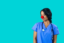 Portrait Of A Smiling Female Doctor Or Nurse Wearing Blue Scrubs Uniform And Red Nose Looking At Camera Isolated On Blue Background