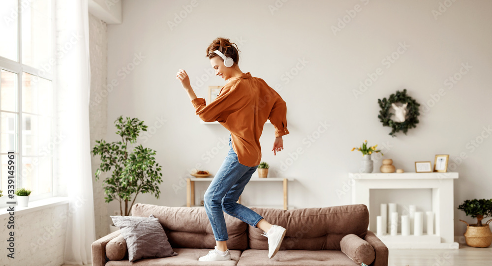 Cheerful woman listening to music and dancing on soft couch at home in day off. - obrazy, fototapety, plakaty 