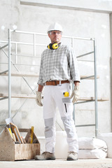 Wall Mural - man construction worker holding tool box wear gloves, hard hat, glasses and hearing protection headphones, at interior building site with scaffolding