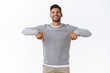 Happy carefree, friendly handsome man with bristle in grey casual sweater, joyfully talking, give advice, recommend promo as pointing bottom, indicate down link, standing white background