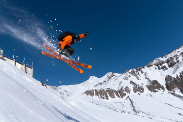 a male athlete skier in an orange trigger makes a jump jump with a grab with flying snow powder agai