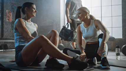 Wall Mural - Two Beautiful Fit Athletic Girls Sit on a Floor of Industrial Loft Gym. They're Happy with their Training Program and Successfully Give a High Five. Strong Masculine Man Walks in the Background.