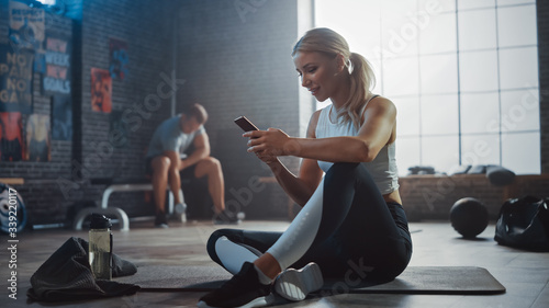 Fotografía Happy and Smiling Beautiful Athletic Young Woman is Using a Smartphone while Sitting on a Floor in a Loft Gym