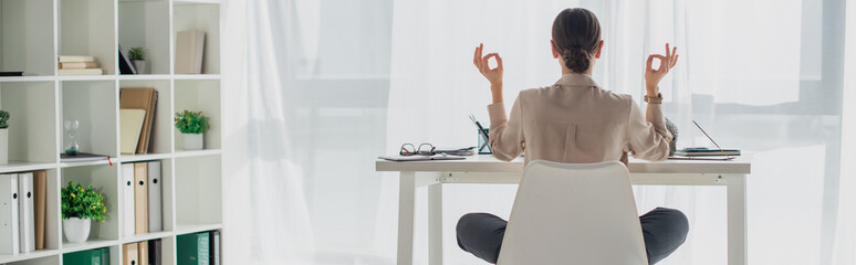 panoramic shot of businesswoman meditating in lotus pose with gyan mudra at workplace with incense s