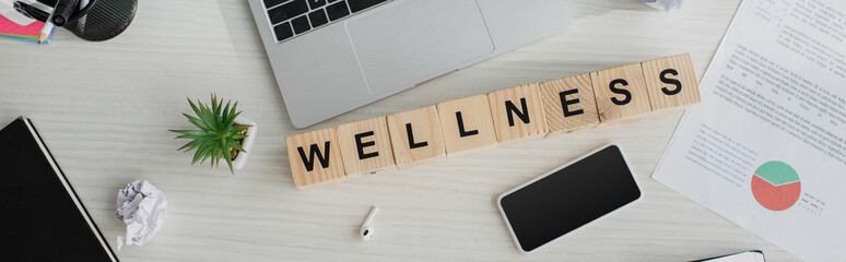 panoramic shot of business workplace with earphones, laptop, smartphone and alphabet cubes with wellness word