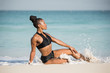 © Paul - Beautiful African American Black female fitness model on the beach wearing black sportswear sitting on the waters edge being splashed by a small wave.
