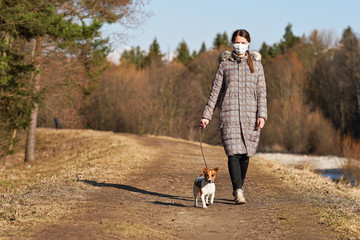 Wall Mural - Young woman in warm jacket, wearing virus face mouth mask, walks her dog on country road. Masks are mandatory   outside home during coronavirus covid-19 outbreak in some countries