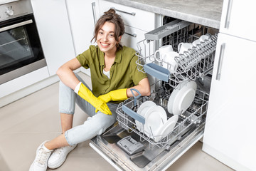 Wall Mural - Portrait of a happy housewife sitting near the dishwasher with clean dishes on the kitchen at home. Easy house work with kitchen appliances concept