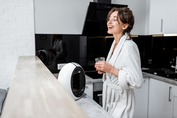 Portrait of a young happy woman in bathrobe standing with coffee on the kitchen with modern appliances at home